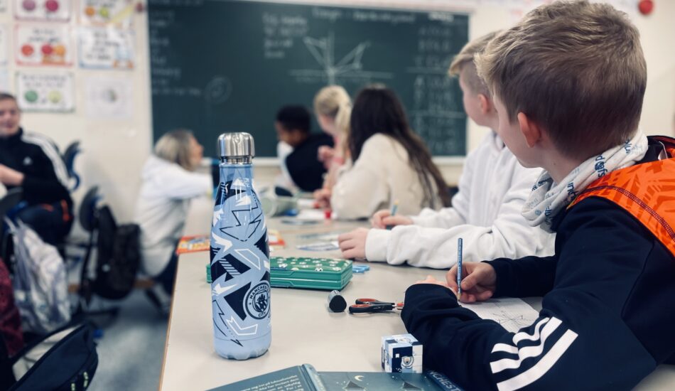 Fotografi. Barn i en klasseromssituasjon. Vi ser barna bakfra, og de ser opp mot tavla.