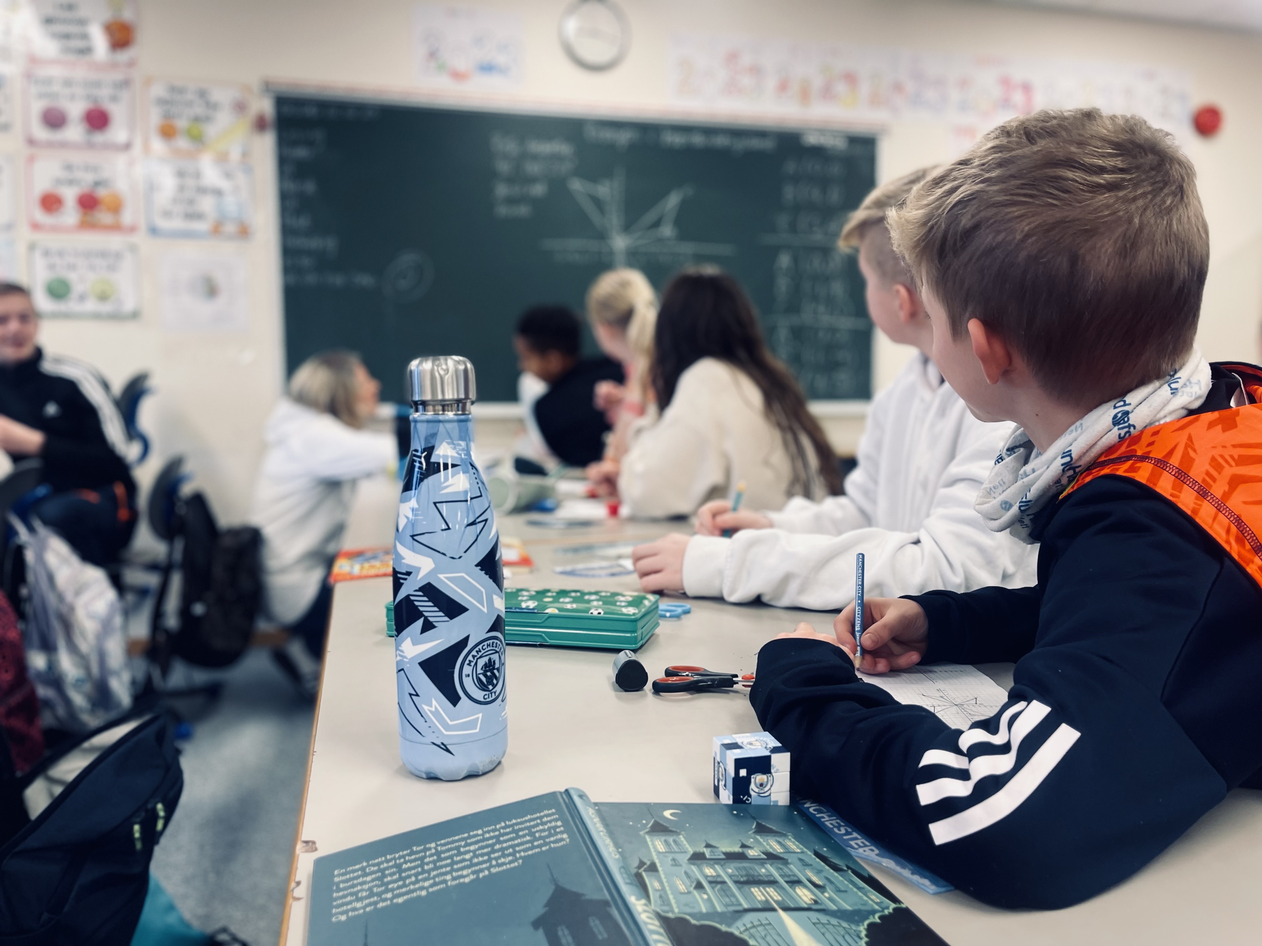 Fotografi. Barn i en klasseromssituasjon. Vi ser barna bakfra, og de ser opp mot tavla.